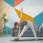 Portrait of cute lady in leggings and top doing yoga at home working out standing on mat exercising. Healthy youth lifestyle and indoor activities concept.