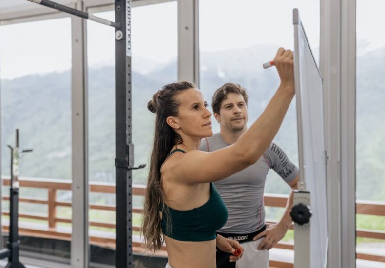 Female fitness coach writes on whiteboard as a male athlete watches in a gym.